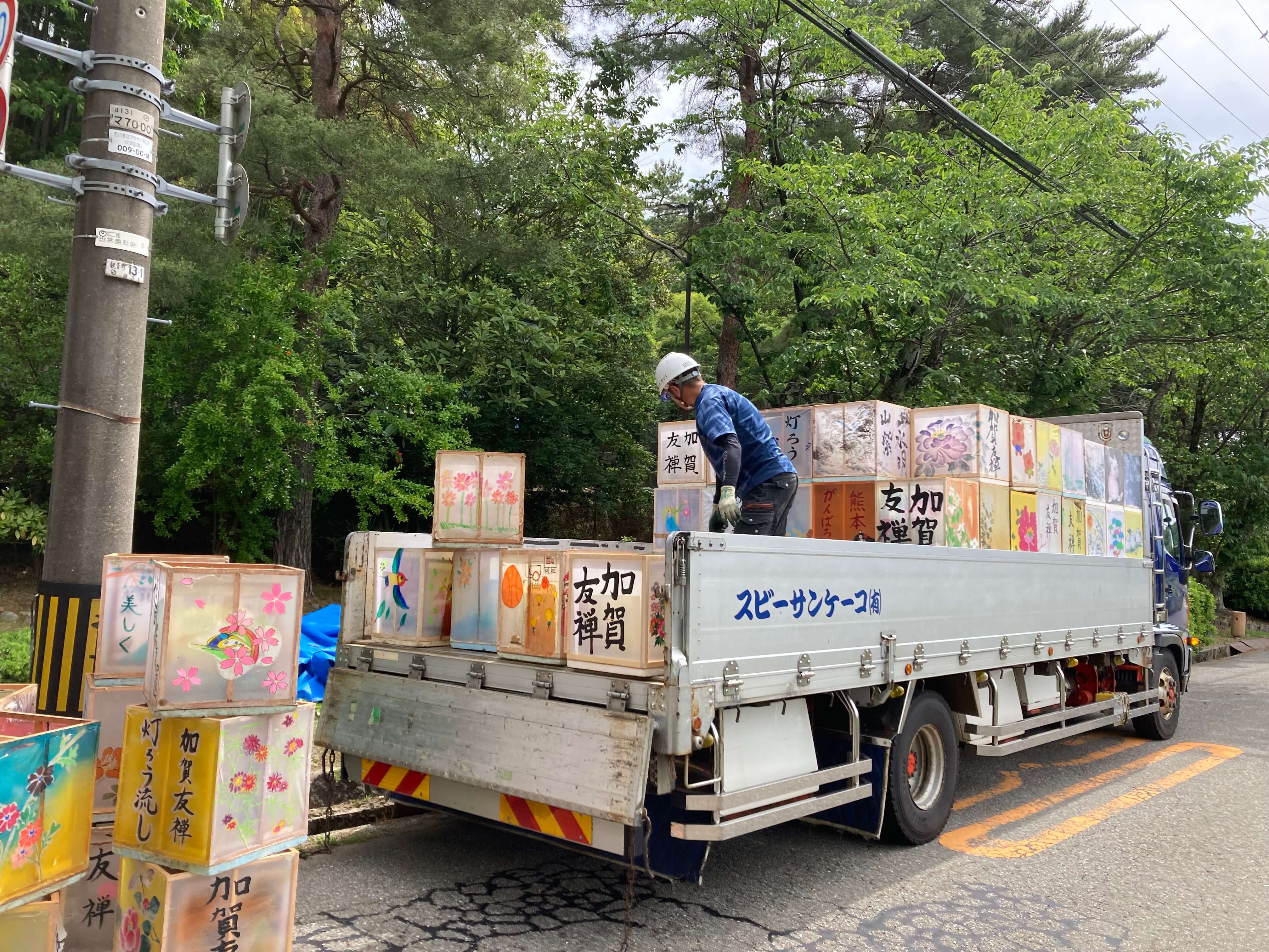 トラックの荷台で提灯や灯籠を運搬している作業員と、道路脇に積み上げられたカラフルな和風の灯籠。日本の祭りやイベントの準備風景。